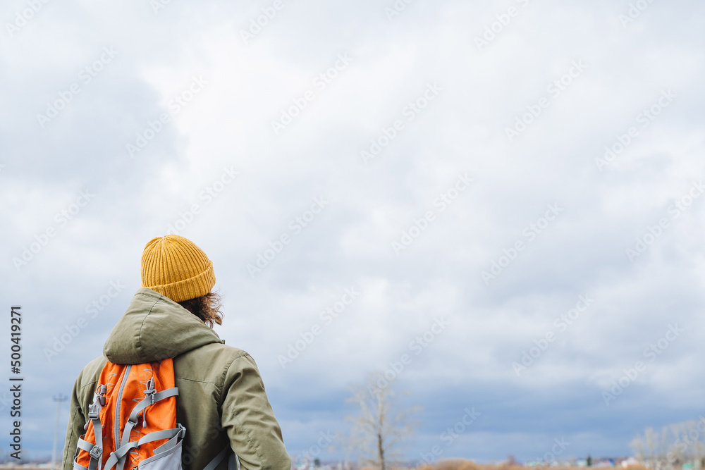 A tourist in a hat with a backpack stands with his back against the sky, a hiking photo, a lonely wanderer, cloudy weather, gray clouds, warm clothes. A tourist in a hat with a backpack stands with his back against the sky, a hiking photo, a lonely wanderer, cloudy weather, gray clouds, warm clothes.