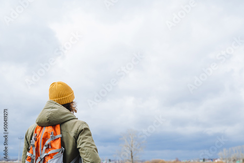 A tourist in a hat with a backpack stands with his back against the sky, a hiking photo, a lonely wanderer, cloudy weather, gray clouds, warm clothes. © Aleksey
