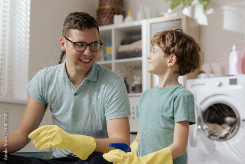 Housekeeping husband and cleaning concept, happy young man in yellow rubber gloves to wipe dust with spray and brush while cleaning on the floor at home with his son.