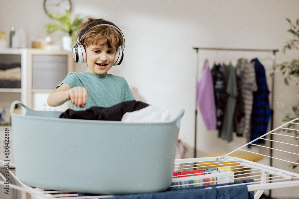 Boy wearing wireless headphones stands on stool in laundry room by fold