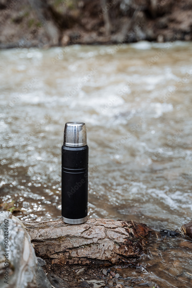 A thermos stands on the background of water, a black vacuum bottle, a thermo mug for hot drinks. A thermos stands on the background of water, a black vacuum bottle, a thermo mug for hot drinks.