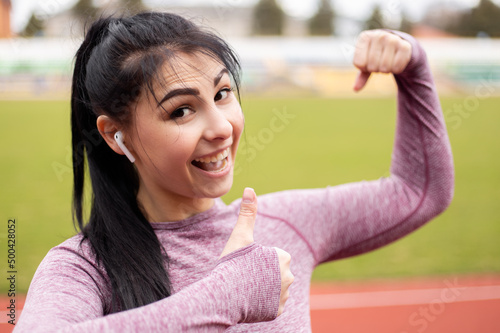 Woman dressed sports suit showing muscles biceps triceps while training doing sport at the stadium treadmill. Athlete posing body, thumb down dislike gesture sign bragging with achievements 