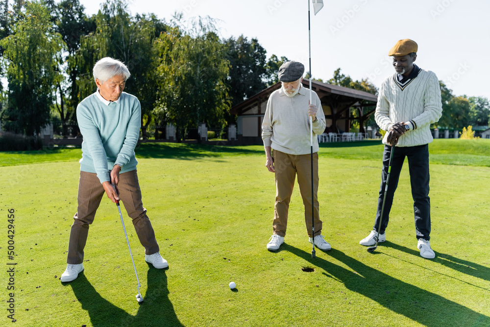 Obraz premium senior man in flat cap holding flag stick near hole and multiethnic friends.