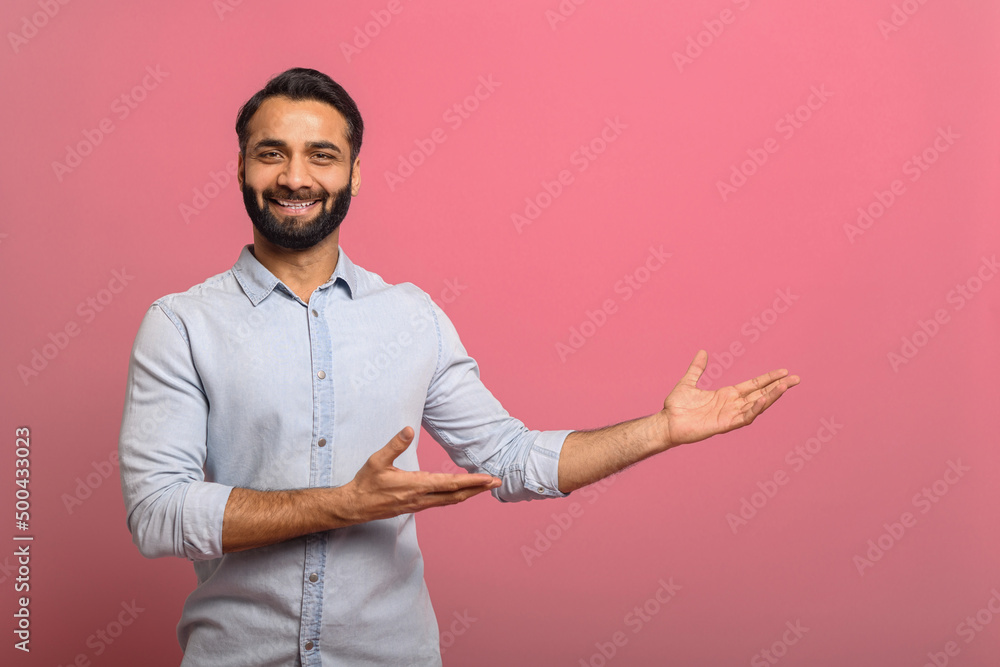 Handsome Indian guy points palms at empty copy space isolated on pink background. Cheerful multiracial bearded man in casual jeans shirt presenting novelty, advertising concept Handsome Indian guy points palms at empty copy space isolated on pink background. Cheerful multiracial bearded man in casual jeans shirt presenting novelty, advertising concept