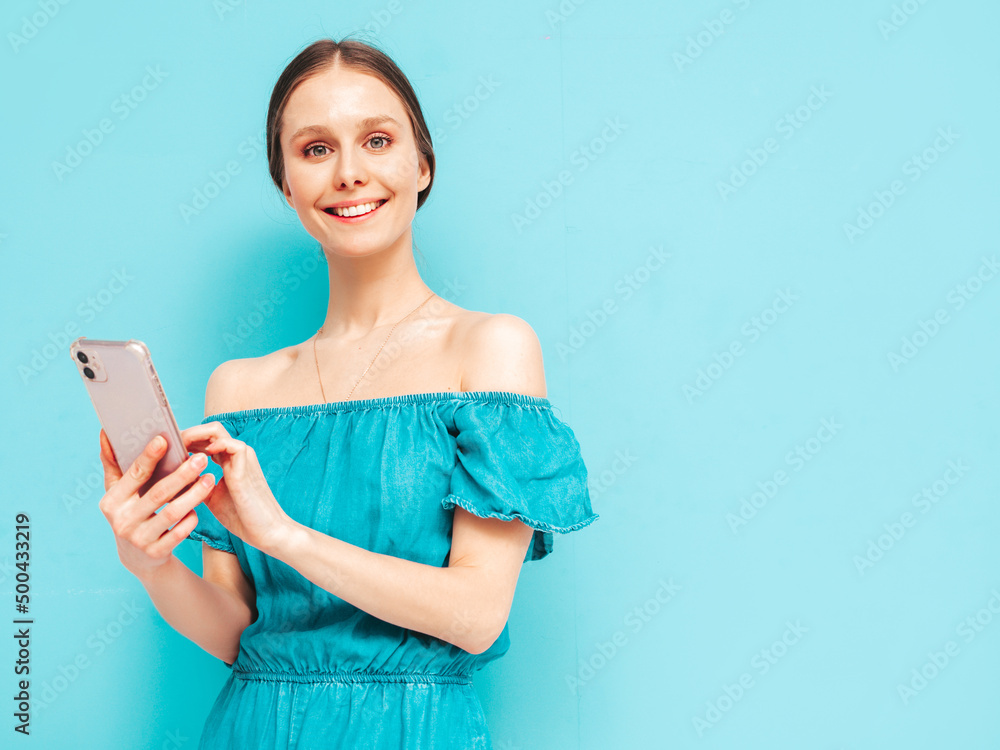Young beautiful smiling female in trendy summer blue overalls. Sexy carefree woman posing near wall in studio. Positive brunette model looking at cellphone screen. Holding smartphone and using apps Young beautiful smiling female in trendy summer blue overalls. Sexy carefree woman posing near wall in studio. Positive brunette model looking at cellphone screen. Holding smartphone and using apps