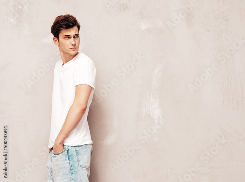 Portrait of handsome confident stylish hipster lambersexual model.Man dressed in white t-shirt and jeans. Fashion male posing in studio near grey wall