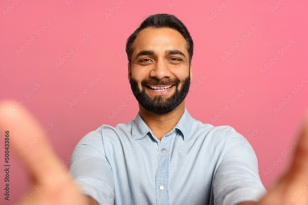 Portrait of bearded Indian handsome man taking selfie, looking at camera POV, multiracial brown-haired guy recording himself. Indoor studio shot isolated on pink background