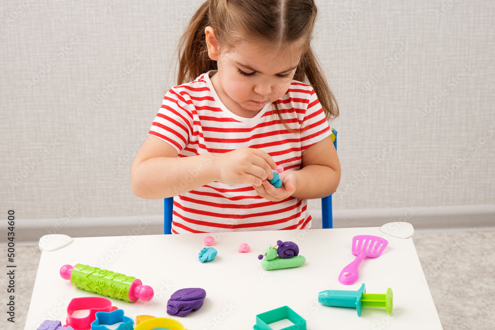 little girl in bright striped T-shirt playing with plasticine, making snails on white table, home educational games little girl in bright striped T-shirt playing with plasticine, making snails on white table, home educational games