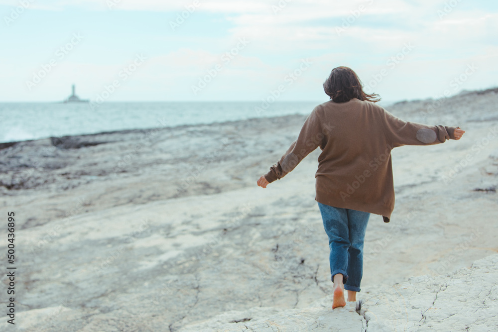 woman walking by rocky sea beach in wet jeans lighthouse on background
