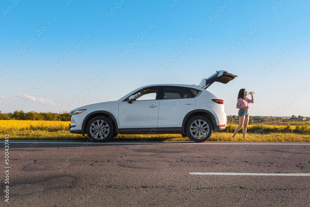 woman drinking water standing near car trunk on sunset at roadside