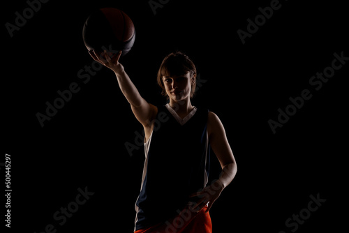 Female basketball player. Beautiful girl holding ball. Side lit silhouette studio portrait against black background © Nikola Spasenoski
