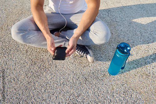 Top view of a sportsman wearing earphones sitting on the city bridge and checking his phone while relaxing after morning cardio workout and using a fitness tracking mobile application