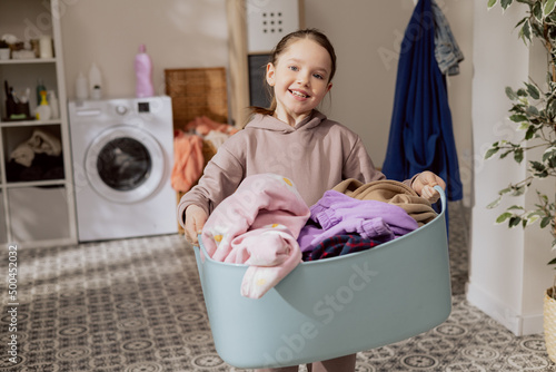 Smiling pretty girl stands in the middle of the bathroom, laundry room, holding a large bowl filled with colorful clothes in her hands, daughter helps her mother with housework.