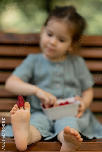 Baby girl eating raspberry on picnic and put on feet.