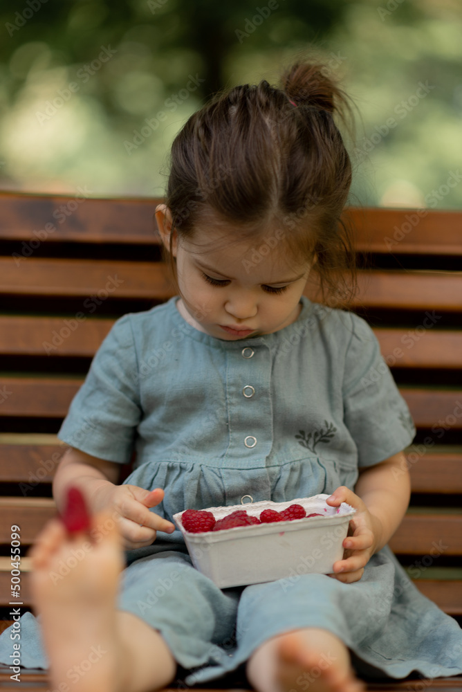 Baby girl eating raspberry on picnic and put on feet.