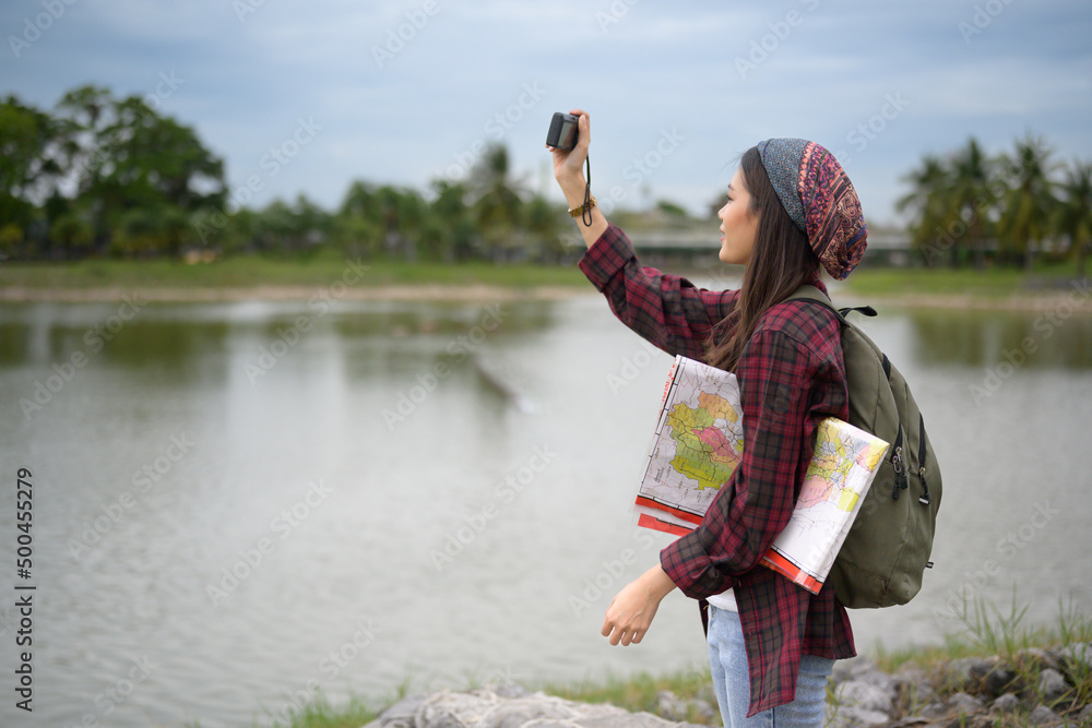 Woman traveler hold camera and taking a photo in the park during vacation on blue sky background.Asian hipster young girl backpack tourist hand holding a map for search and plan her trip.