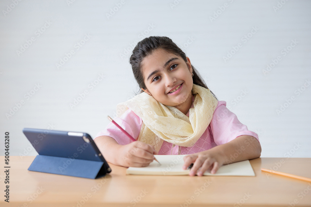 portrait cute Indian girl writing on notebook and online learning class from tablet, education concept portrait cute Indian girl writing on notebook and online learning class from tablet, education concept