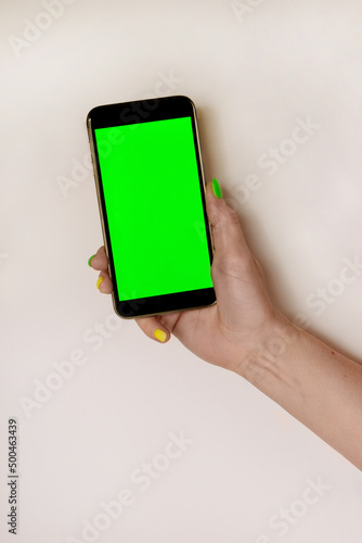 Close-up of a female hand holding a mobile phone with a vertical green screen on a light table. Vertical photography