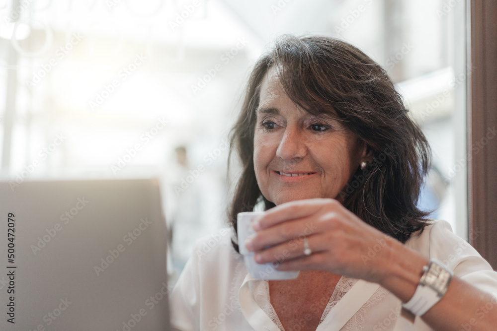 Smiling woman sitting in cafeteria holding coffee mug and working on laptop. Businesswoman checking email on laptop. Beautiful middle aged woman and using laptop at cafe while drinking a cup of tea. Smiling woman sitting in cafeteria holding coffee mug and working on laptop. Businesswoman checking email on laptop. Beautiful middle aged woman and using laptop at cafe while drinking a cup of tea.