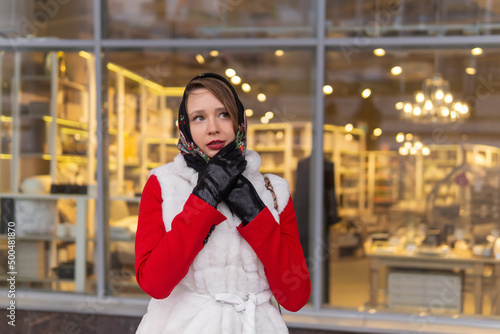 young woman in a headscarf in cold weather stands outside against the background of a blurry bright shop window