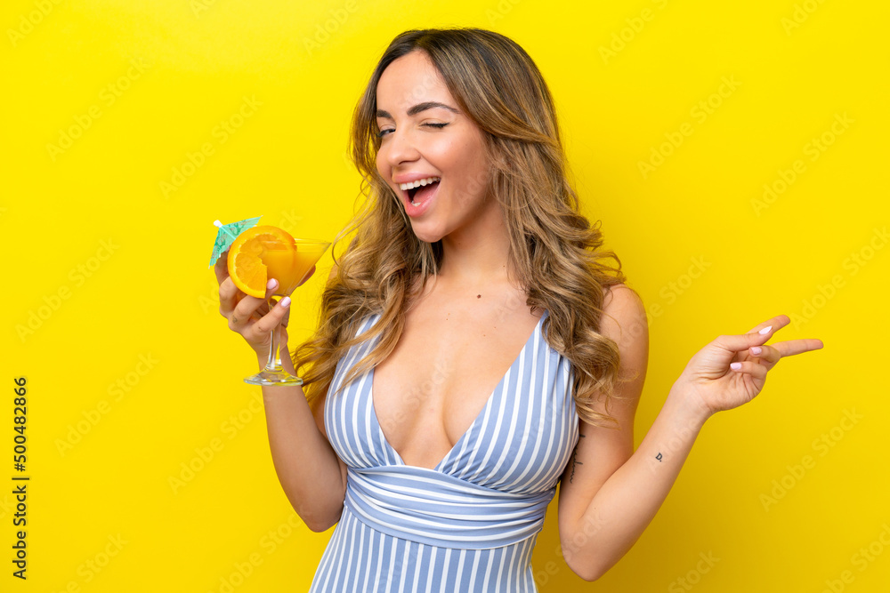 Young caucasian woman isolated on yellow background in swimsuit and holding a cocktail