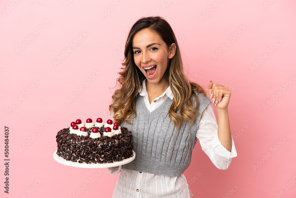 Young woman holding birthday cake over isolated pink background celebrating a victory in winner position Young woman holding birthday cake over isolated pink background celebrating a victory in winner position