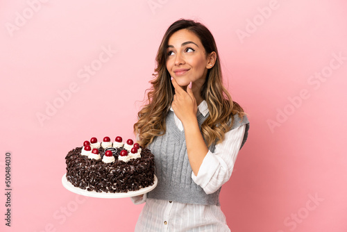 Young woman holding birthday cake over isolated pink background looking up while smiling