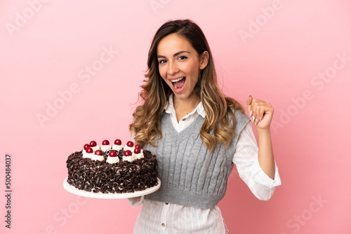 Young woman holding birthday cake over isolated pink background celebrating a victory in winner position