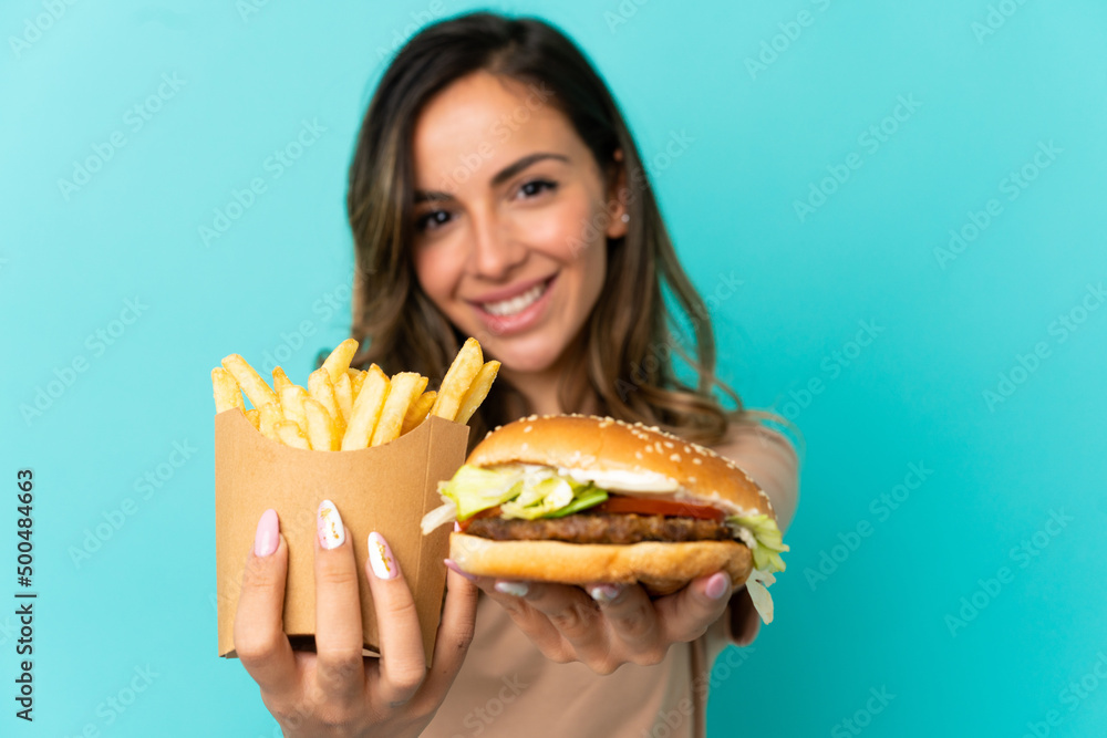 Young woman holding fried chips and burger over isolated background Young woman holding fried chips and burger over isolated background