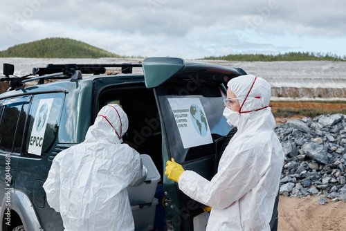 Rear view of polluttion scientists in protective workwear and masks opening car trunk door while getting tool out of container on abandoned territory