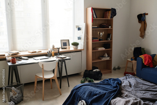 Modern minimalistic students room with awards on shelves, unmade bed with blanket and office supplies on desk