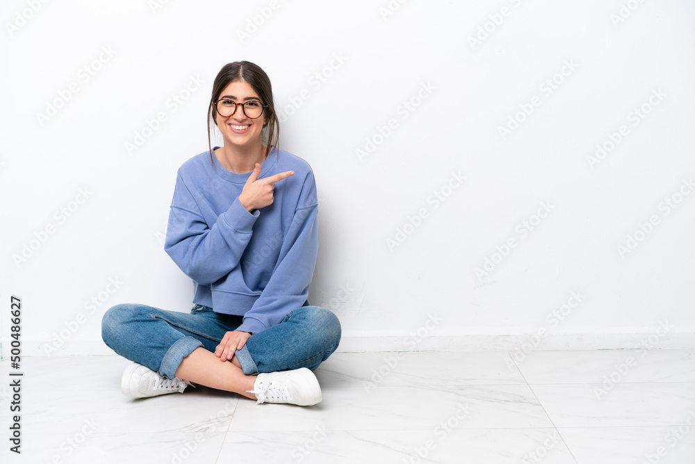 Young caucasian woman sitting on the floor isolated on white background pointing finger to the side Young caucasian woman sitting on the floor isolated on white background pointing finger to the side