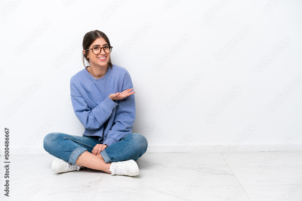 Young caucasian woman sitting on the floor isolated on white background presenting an idea while looking smiling towards
