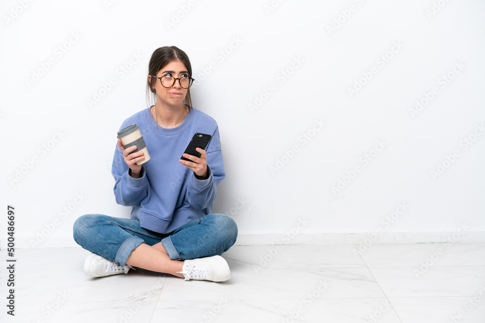 Young caucasian woman sitting on the floor isolated on white background holding coffee to take away and a mobile while thinking something Young caucasian woman sitting on the floor isolated on white background holding coffee to take away and a mobile while thinking something