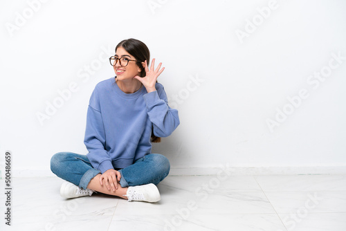 Young caucasian woman sitting on the floor isolated on white background listening to something by putting hand on the ear © luismolinero