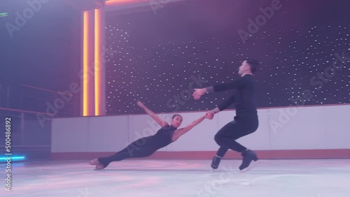 Pair of figure skaters rotating on ice rink,woman goes into death spiral,hovers in air parallel to ice her legs in split, man holds her by hands. Figure skating lift,slow motion support,distant shot.