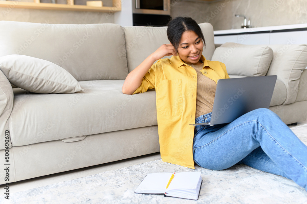 Charming and calm African-American woman sits on the floor in cozy living room with a laptop, enjoys lazy weekend in relaxed atmosphere, working remotely from home, chatting or studying online Charming and calm African-American woman sits on the floor in cozy living room with a laptop, enjoys lazy weekend in relaxed atmosphere, working remotely from home, chatting or studying online
