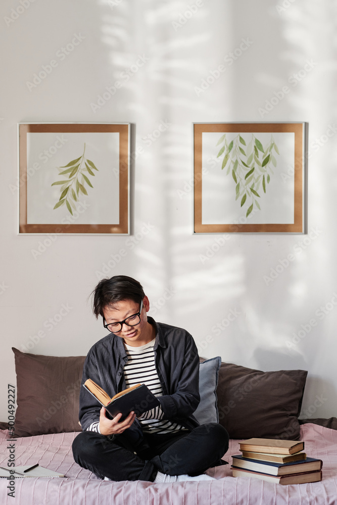Smiling Asian student boy in shirt sitting with crossed legs and reading textbook in cozy room