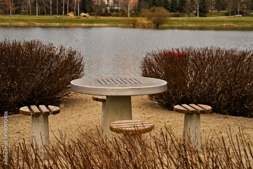 Parkowy stolik do szachów ( i nie tylko ) wraz z siedziskami , wśród krzewów ozdobnych , nad stawem wczesną wiosną . 