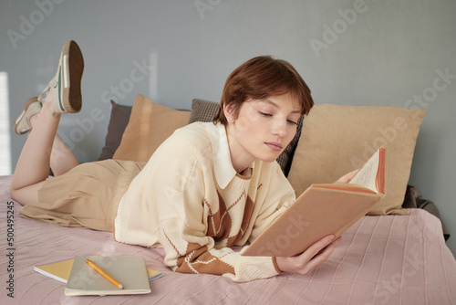 Serious short-haired girl in students uniform lying on bed and reading textbook while prepaing for classes photo