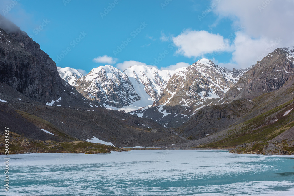 Atmospheric highland landscape with frozen alpine lake and high snowy mountains. Awesome scenery with icy mountain lake on background of large snow mountains under clouds. Scenic view to ice lake.