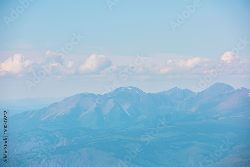 Minimalist aerial landscape with forest hills and high mountain range in sunlight and haze at changeable weather. Minimal alpine scenery with sunlit mountain tops silhouettes in mist under cloudy sky.