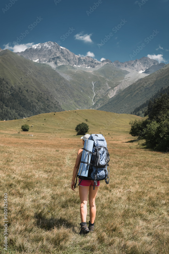 Tourist with large backpacks climbs the grassy slope overlooking the snow-capped peaks in the Caucasus Mountains Georgia Svaneti