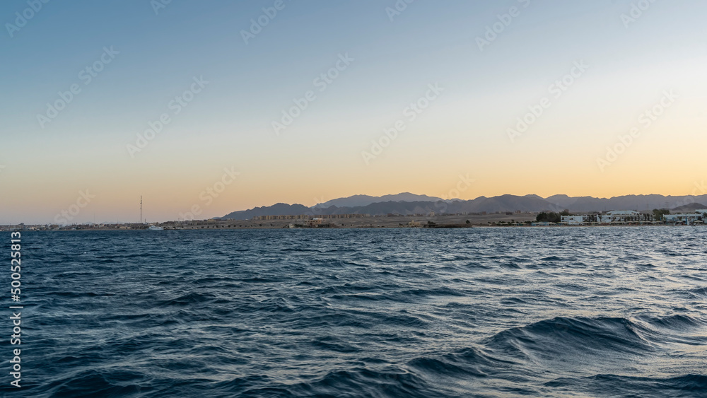Evening in Egypt. A picturesque mountain range against a blue-orange sky. Hotel houses are visible on the shore. Waves on the surface of the Red Sea. Safaga