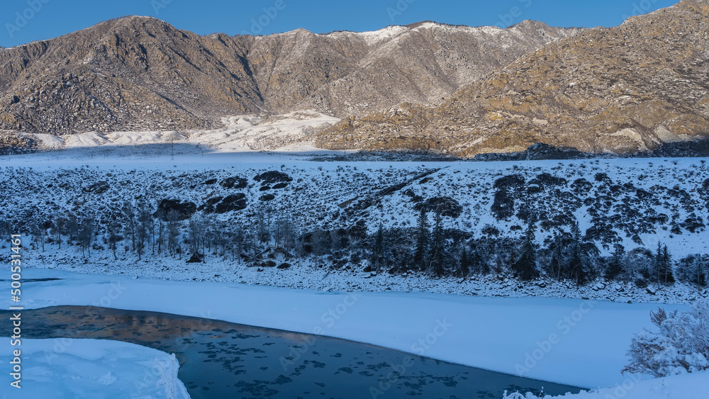 Turquoise ice-free river between the icy shores. Thawed ice floes are visible in the riverbed. There are coniferous trees on the snow-covered shores. The mountain range is illuminated by the sun