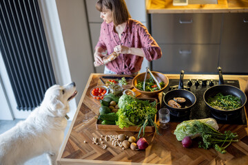 Young housewife makes a salad while standing with her adorable white dog in the kitchen. Table with lots of fresh green food ingredients. Healthy eating and leisure time with pet