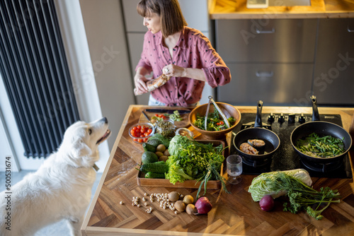Young housewife makes a salad while standing with her adorable white dog in the kitchen. Table with lots of fresh green food ingredients. Healthy eating and leisure time with pet