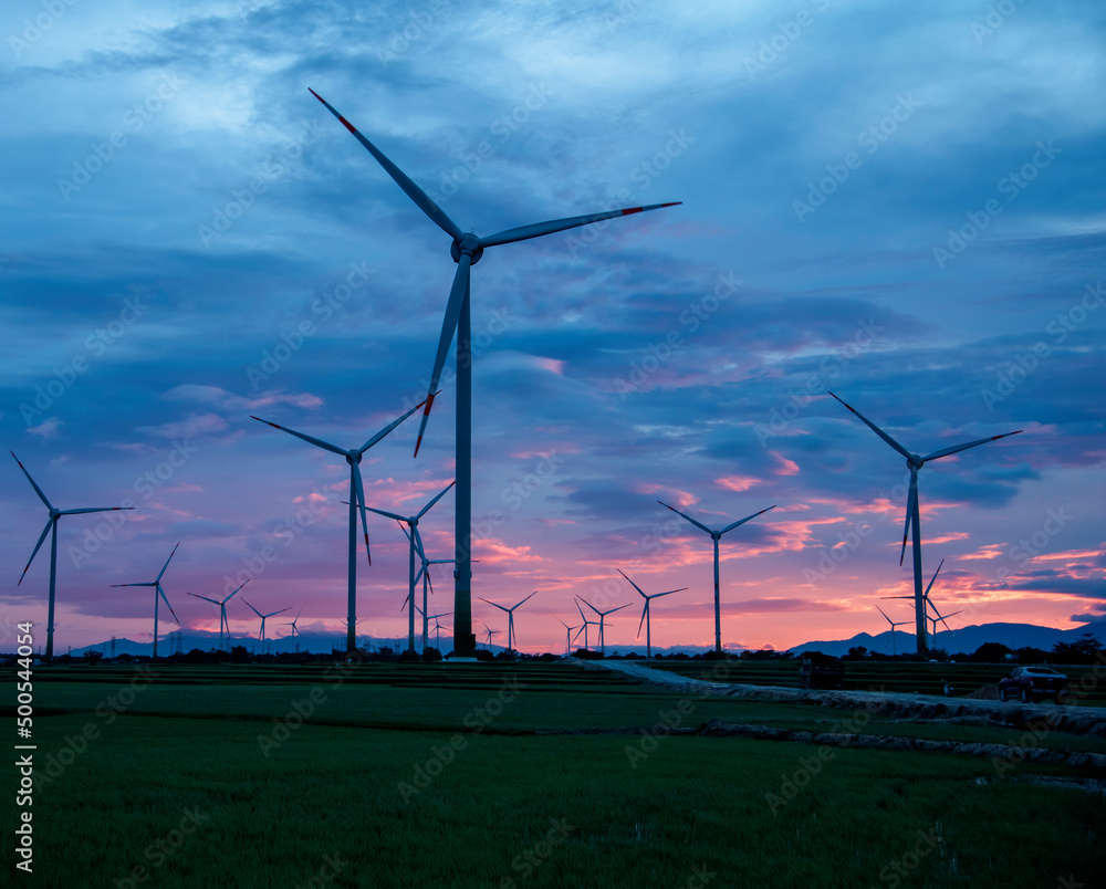 Wind farm or wind park, with high wind turbines in the setting sun for generation electricity with copy space. Green energy concept to reduce climate change and global warming Wind farm or wind park, with high wind turbines in the setting sun for generation electricity with copy space. Green energy concept to reduce climate change and global warming