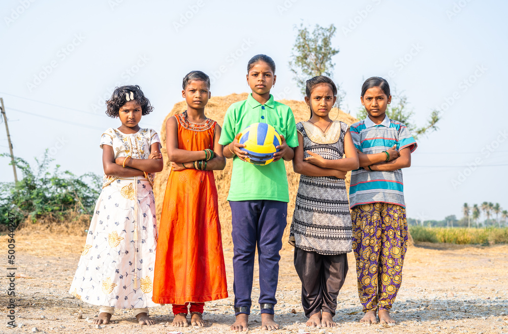 full shot, group of confidently standing Indian village girls football by looking at camera during training - concept of positive emotion, summer training, leisure activities and hobbies