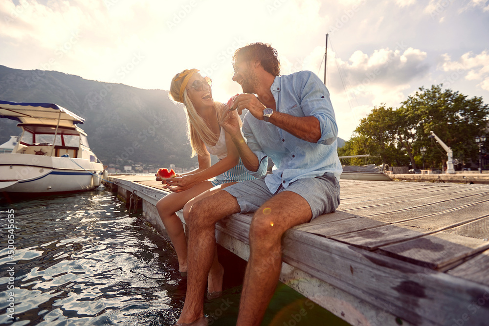 Lovely young couple sharing moments of bliss and eating watermelon sitting on wooden jetty by water. Summertime holiday by seaside. Togetherness, lifestyle, luxury concept. - obrazy, fototapety, plakaty 